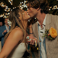 Bride and groom with cake on their faces share a kiss at an outdoor wedding reception. String lights create a festive atmosphere, while guests watch in the background. The groom wears a flower boutonniere.