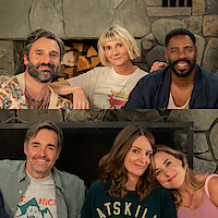 Six adults sitting closely together in front of a stone fireplace, smiling warmly in a cozy living room setting, wearing casual clothes, suggesting a relaxed and friendly gathering indoors.