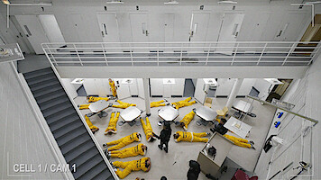Prison common area with multiple inmates in yellow uniforms lying on the floor, viewed from above. White walls, doors, tables, and stairs in a secure facility setting. Surveillance camera footage.