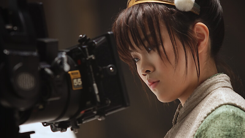 Miyako Cech as Toph on a film set, focused intently on a professional movie camera during a scene, with soft lighting and a blurred studio background.