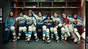 A group of young hockey players in blue and white uniforms and one in red sit together on a locker room bench, posing and smiling for a team photo, hockey gear and sticks by their sides.