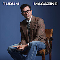 Dan Levy poses against a blue backdrop, wearing a checked blazer.