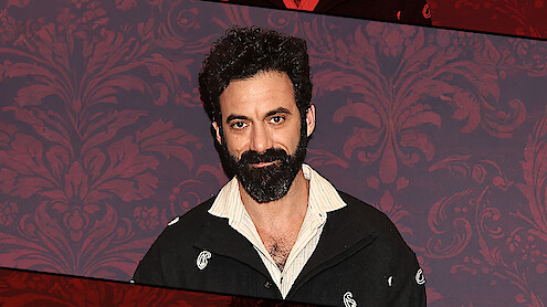 A man with dark curly hair and a full beard stands indoors against a patterned red wall, wearing a black jacket with a partially open collared shirt, posing for a portrait with a neutral expression.