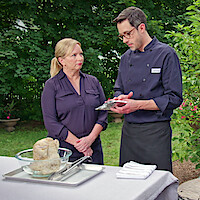 Two people in chef attire stand outside by a table set with cooking ingredients and utensils, discussing a recipe in a lush garden setting.