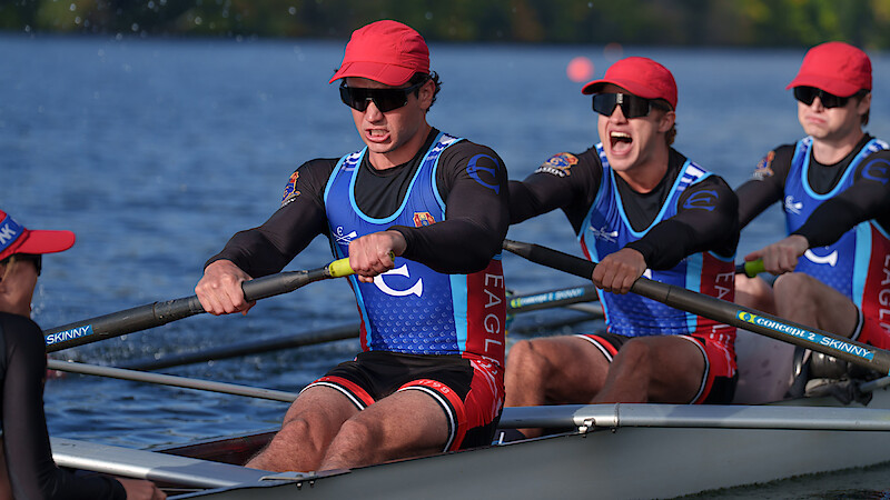 Rowers in matching uniforms and red hats compete in a crew race on a calm river, focused and working together in an outdoor setting with greenery in the background.