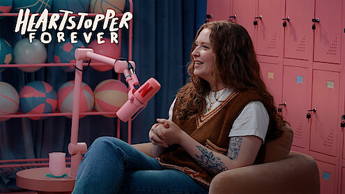 Alice Oseman sits in a cozy chair speaking into a pink microphone, with pink lockers and sports balls in the background, and "Heartstopper Forever" logo displayed in the corner.