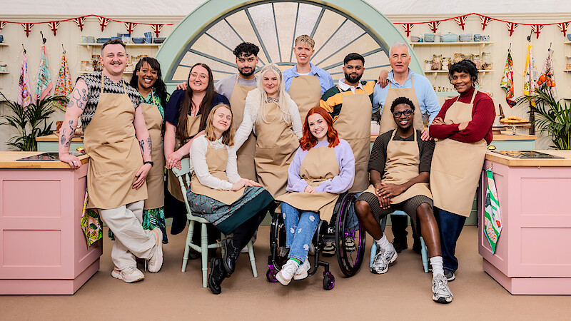 A diverse group of twelve people in aprons pose inside a colorful baking tent with pink counters, plants, and festive bunting, smiling for a group photo.