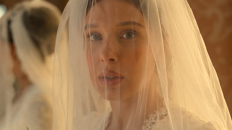 A bride in a white lace dress and veil looks solemnly forward in soft, warm lighting, with a blurred reflection behind her, possibly in a dressing room or bridal suite.