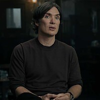 A man with medium-length dark hair wearing a brown sweater sits indoors in a dimly lit room, hands folded, with a blurred background suggesting a cozy or intimate setting.