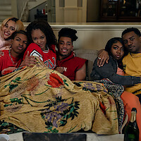 Six friends sitting close together on a couch in a cozy living room, wrapped in a floral blanket, relaxed and enjoying each other’s company. The setting is warm, inviting, and casual, with drinks on the table in front of them.