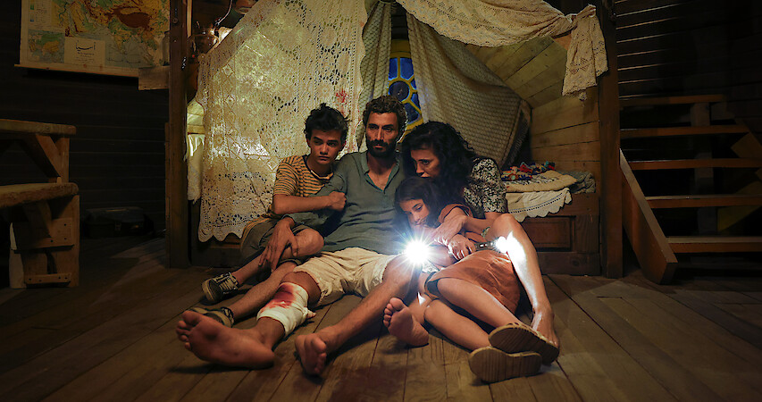 Riman Al Rafeea, Ziad Bakri, Zain Al Rafeea, and Nadine Labaki huddle together in a small room on a wood floor in the dark in an image from the 2025 film 'The Sand Castle.'