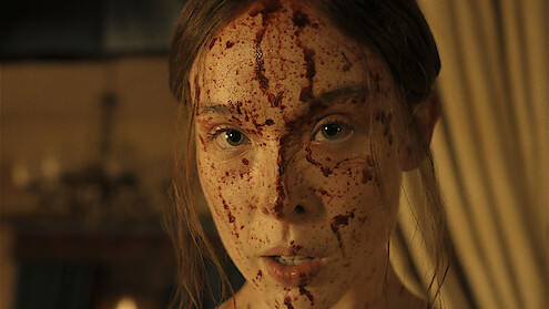 A woman with blood splattered on her face stands indoors in a dimly lit room with curtains and a chandelier visible in the background, looking directly at the camera with an intense expression.