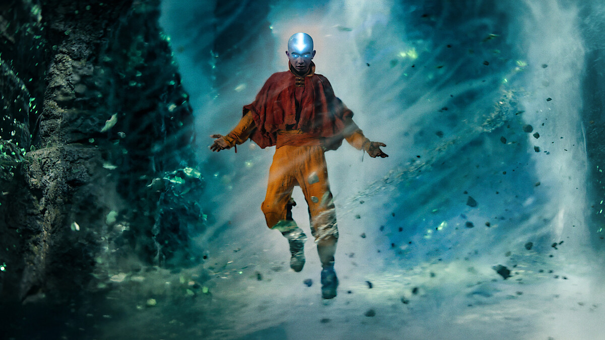 Gordon Cormier as Aang in traditional orange and red robes hovers in the air with glowing light on their head, surrounded by swirling wind and floating debris in a mystical cave or cavern environment.