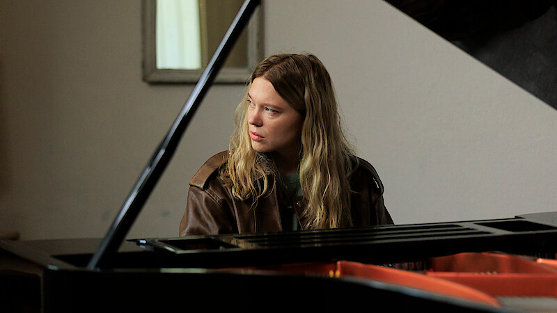 A person with long hair and a brown jacket sits at a grand piano indoors, looking to the side. The setting is softly lit with a neutral wall and a mirror in the background.