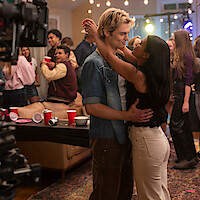 A young couple dances closely at a lively house party with friends in the background, red cups on a table, string lights overhead, and a film crew camera partially visible, capturing the casual atmosphere.