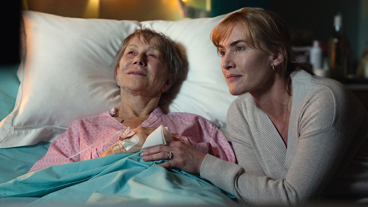 An elderly woman lies in a hospital bed smiling, holding hands with a comforting visitor. 
