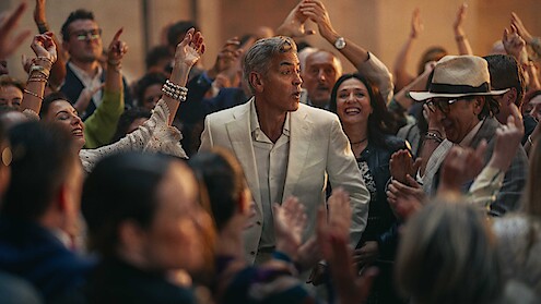 A man in a white suit stands surrounded by a lively crowd at a festive gathering, with smiling faces and raised hands creating a joyful, celebratory atmosphere.