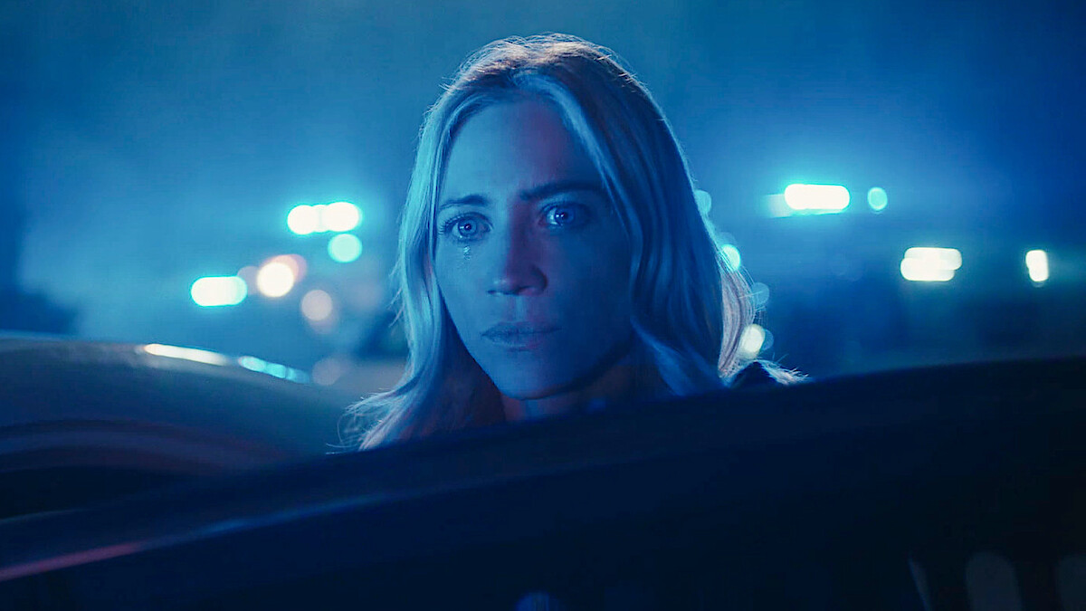 A woman with blonde hair looks serious under blue emergency vehicle lights at night, partially seen through a car window, suggesting a tense or dramatic situation involving law enforcement.