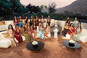 A group of women in elegant dresses sit and stand on white sofas in an outdoor lounge setting at sunset, with mountains, trees, string lights, and round tables with flowers in the background.