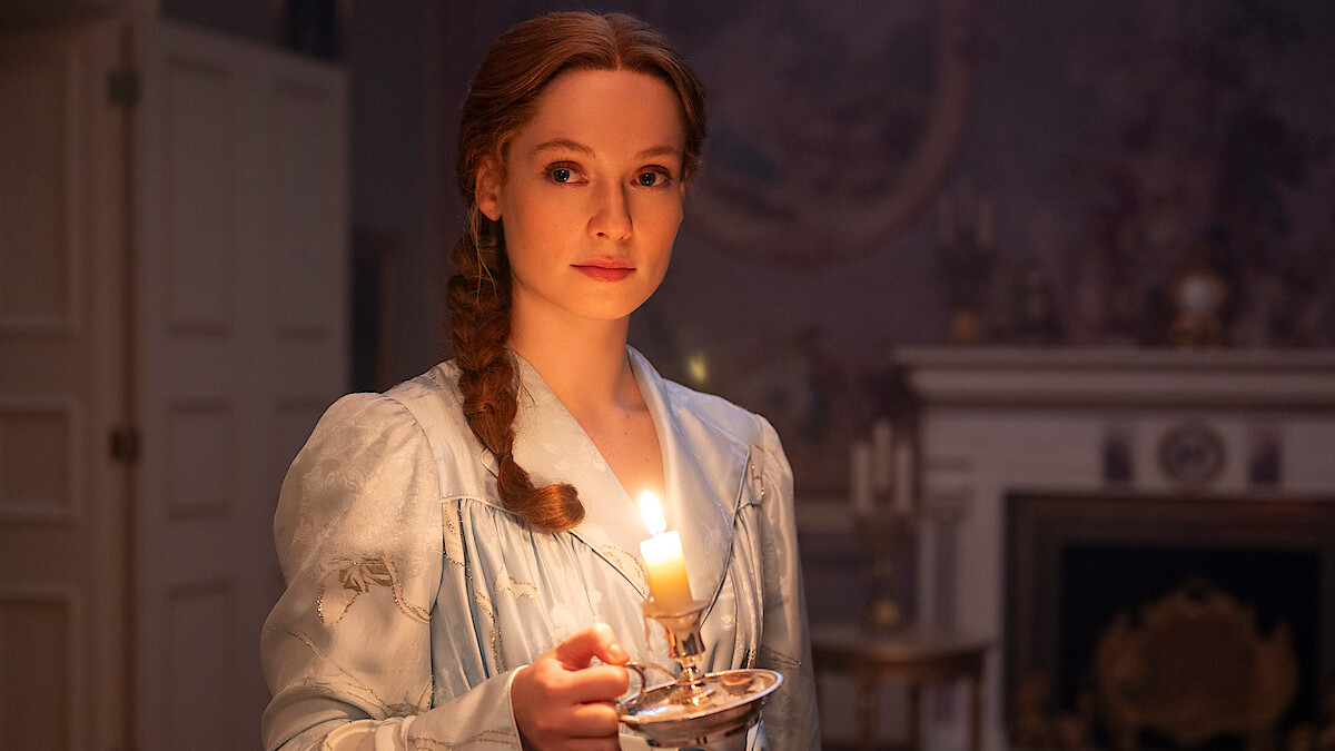 A woman in period clothing holds a lit candle in a dimly lit, vintage-style room with a fireplace and decorative items in the background.