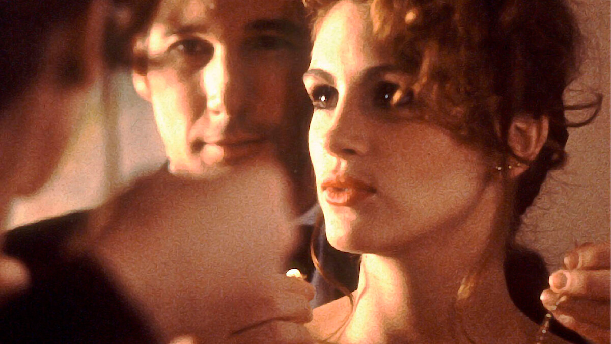 A man and woman stand closely together in a softly lit indoor setting, with the man carefully placing a necklace around the woman's neck, both reflected in a mirror, creating an intimate and elegant atmosphere.