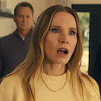 A surprised woman in a cozy indoor setting with soft light, wearing a cream sweater and gold necklace, mouth open in shock, with a man blurred in the background, suggesting a dramatic or emotional moment.