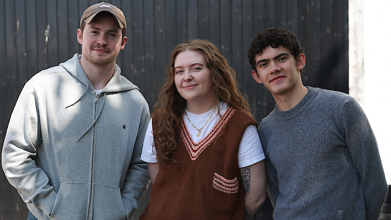 Three young adults standing side by side outdoors in front of a dark wooden wall, smiling at the camera. Casual clothing, natural lighting, and a relaxed atmosphere.
