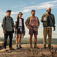 Four hikers in outdoor gear stand on a rocky hilltop with forested landscape and blue sky in the background, prepared for a nature trek or adventure.
