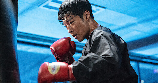 In a blue-lit room, a man wearing a black jacket and red boxing gloves prepares to hit a punching bag.