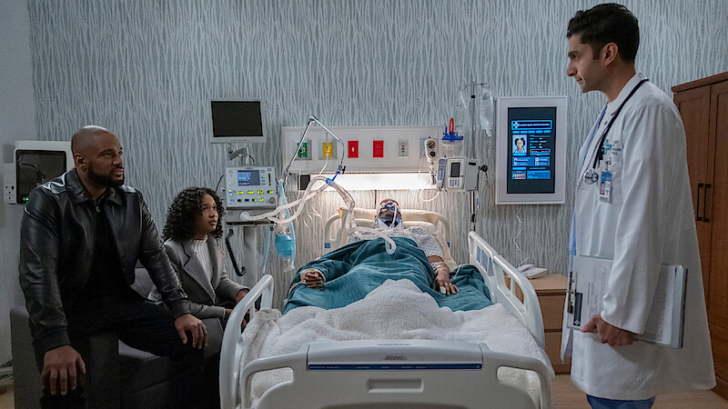 A patient lies in a hospital bed connected to medical equipment, with a man and woman seated nearby and a doctor standing at the bedside in a modern hospital room.