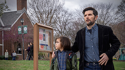 A man and child stand outside near a community notice board with a "Have You Seen Me?" poster. They look alert or concerned. The setting is a grassy area by a brick building with people and leafless trees in the background.