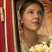 Bride in lace dress and veil holding bouquet, looking thoughtfully out a carriage window with warm light and ornate gold and red details in the background.