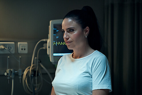 A woman nurse stands smiling conspiratorially in front of an EKG monitor