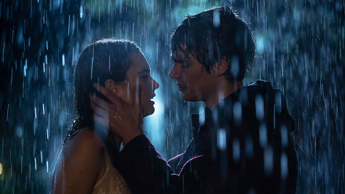 Two people stand close together in the rain at night, gazing into each other's eyes as one gently holds the other’s face, surrounded by falling water and dramatic lighting, creating a romantic and emotional atmosphere.