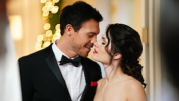 A man in a tuxedo and a woman in a strapless dress stand close, about to kiss, in a warmly lit indoor setting with blurred lights and a festive atmosphere in the background.