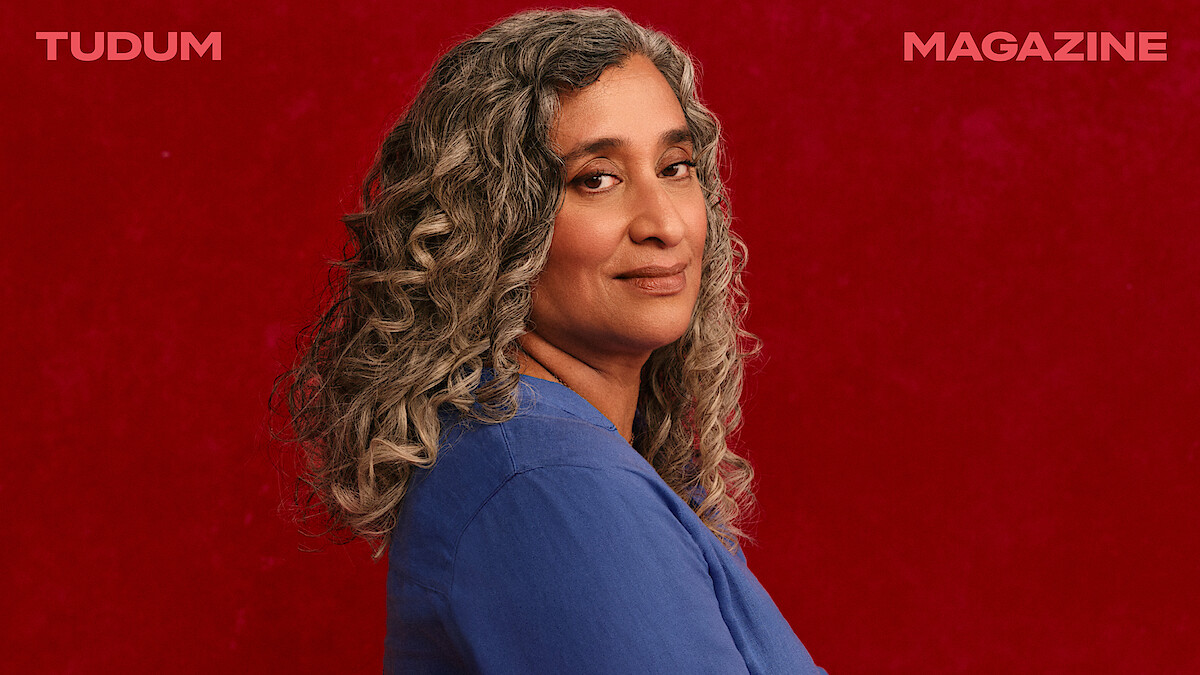 Geeta Gandbhir, director of The Perfect Neighbor, stands in front of a rich red backdrop with beautifully set curls in her graying hair, head turned toward the camera with a confident expression on her face.