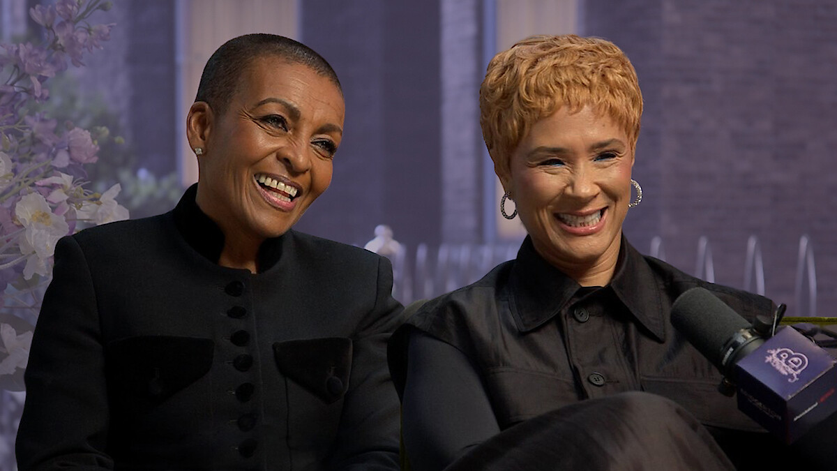 Two women smiling during an interview, seated indoors with a floral arrangement and large windows in the background, both wearing black outfits, with a microphone labeled “20” visible in the foreground.