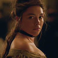 Woman with wavy hair in a period dress and choker necklace looks over her shoulder in a dimly lit, formal setting with blurred people in the background.
