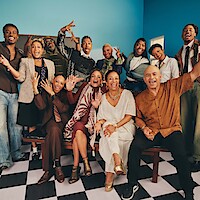 A joyful group of people posing together indoors on a checkered floor, with bright expressions and colorful, stylish clothing against a blue wall, creating a lively and celebratory atmosphere.