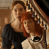 Two women in vintage clothing stand closely together, peering out from behind ornate curtains in an elegant, softly lit room with classic decor and floral arrangements, suggesting a historical or period drama setting.