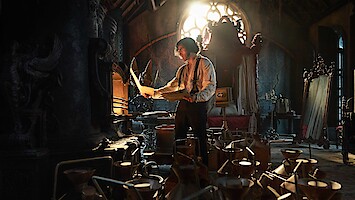 A man studies papers in a dark, ornate room filled with scientific equipment and warm, dramatic light streaming through a stained-glass window, creating a mysterious and intense mood.