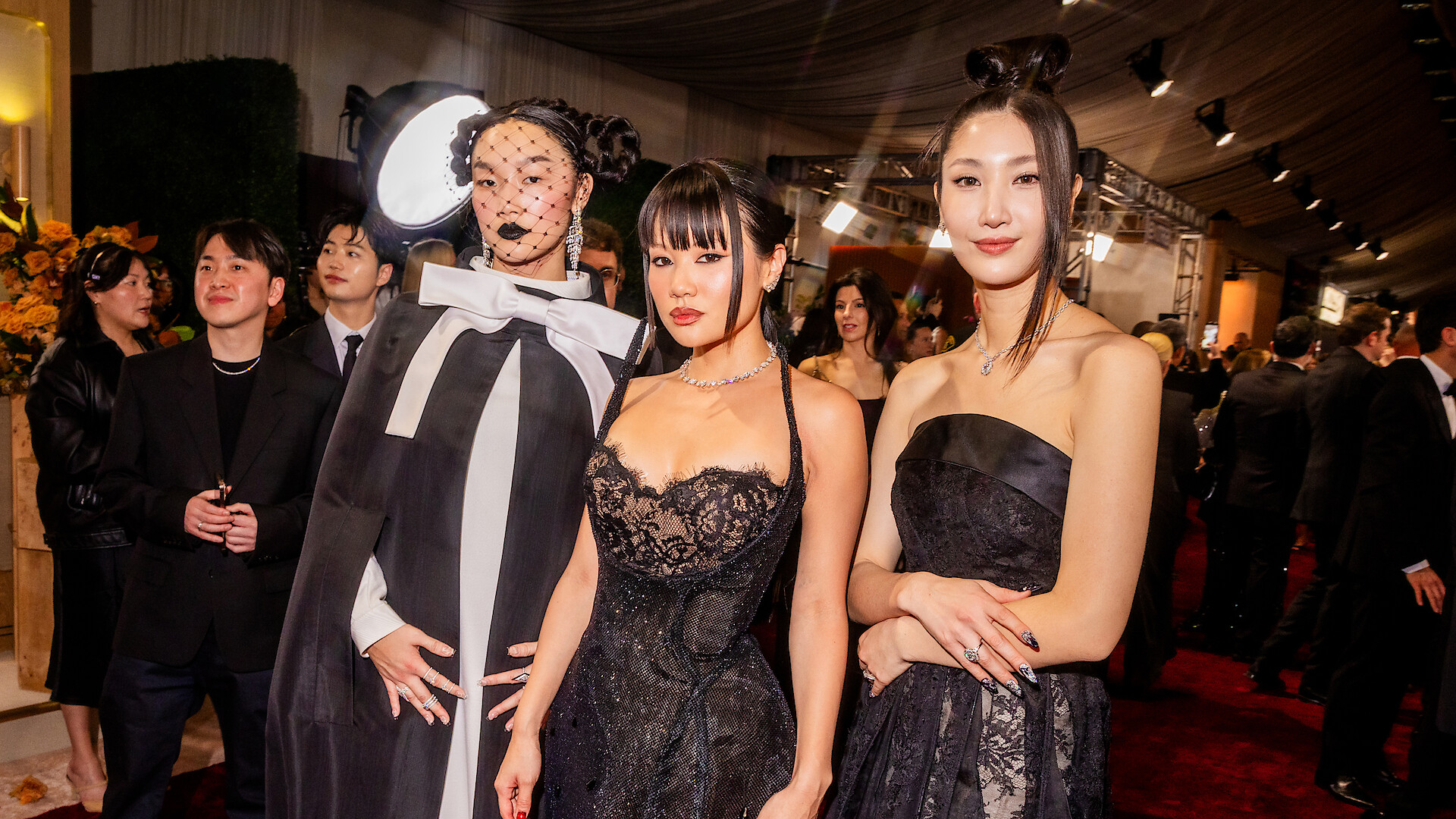 Three people in elegant black and white formal attire pose together at a glamorous indoor event with a red carpet, stylish decor, and other well-dressed guests in the background.
