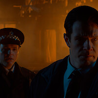 Two serious men, one in a police uniform and another in a suit, stand in a dimly lit, smoky environment with an orange glow, possibly from a fire or emergency scene at night.