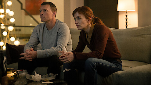 Two people sit on a couch in a dimly lit living room, looking intently at something offscreen. They have takeout containers and drinks on the table, suggesting a casual night at home. Warm lighting creates a cozy and focused atmosphere.