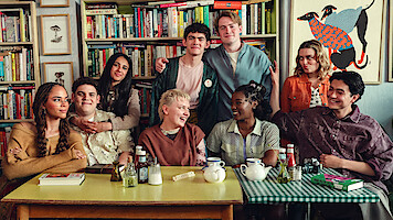 A group of eight young adults sitting and standing around tables with tea and books in a cozy room filled with bookshelves and colorful art, smiling and enjoying each other's company.