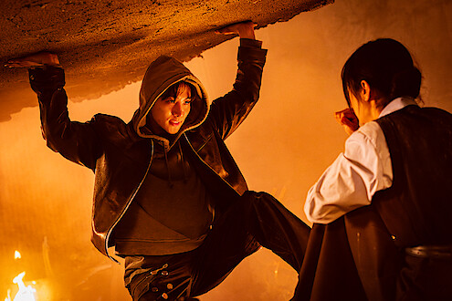 A person in a hoodie holds up a collapsing ceiling in a smoky, fire-lit room while another person crouches nearby, looking at them in concern.