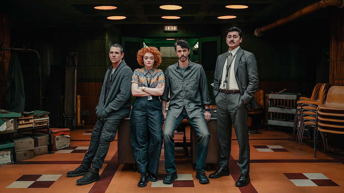 Four people stand in a vintage office with dim lighting and tiled floors, surrounded by stacked chairs, crates, and paperwork, suggesting a detective or investigative setting.