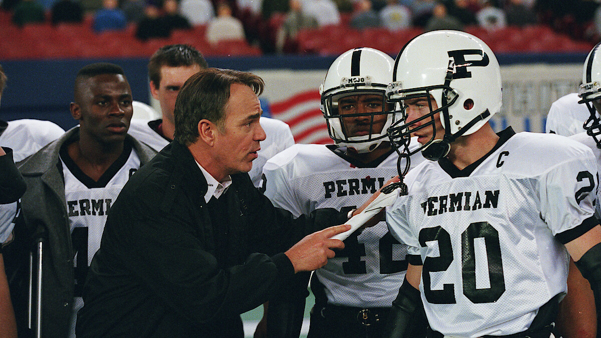 Football coach giving instructions to Permian High School players on the sideline during a game, with players in white uniforms listening intently, inside a stadium with blurred audience in the background.