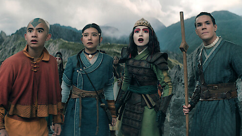 Four young people in fantasy costumes stand closely together outside in rocky mountains, looking ahead with surprised expressions. The setting is dramatic with cloudy skies and rugged terrain behind them.
