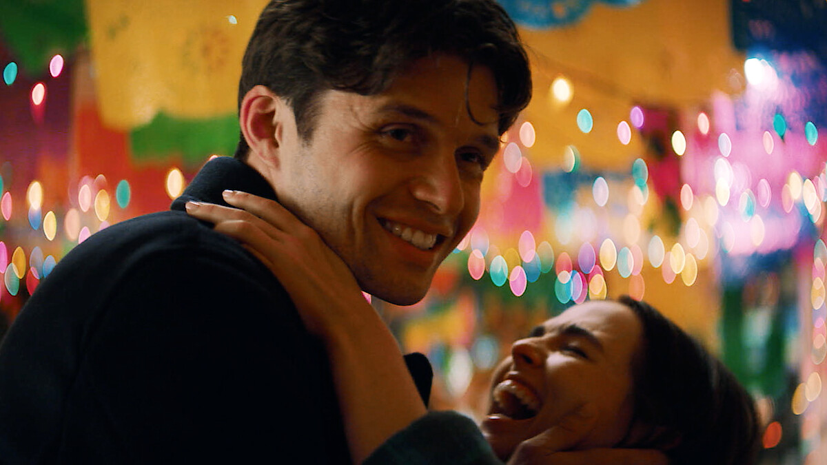 A smiling man embraces a laughing woman at a festive indoor party decorated with colorful lights and banners, creating a joyful, lively atmosphere.
